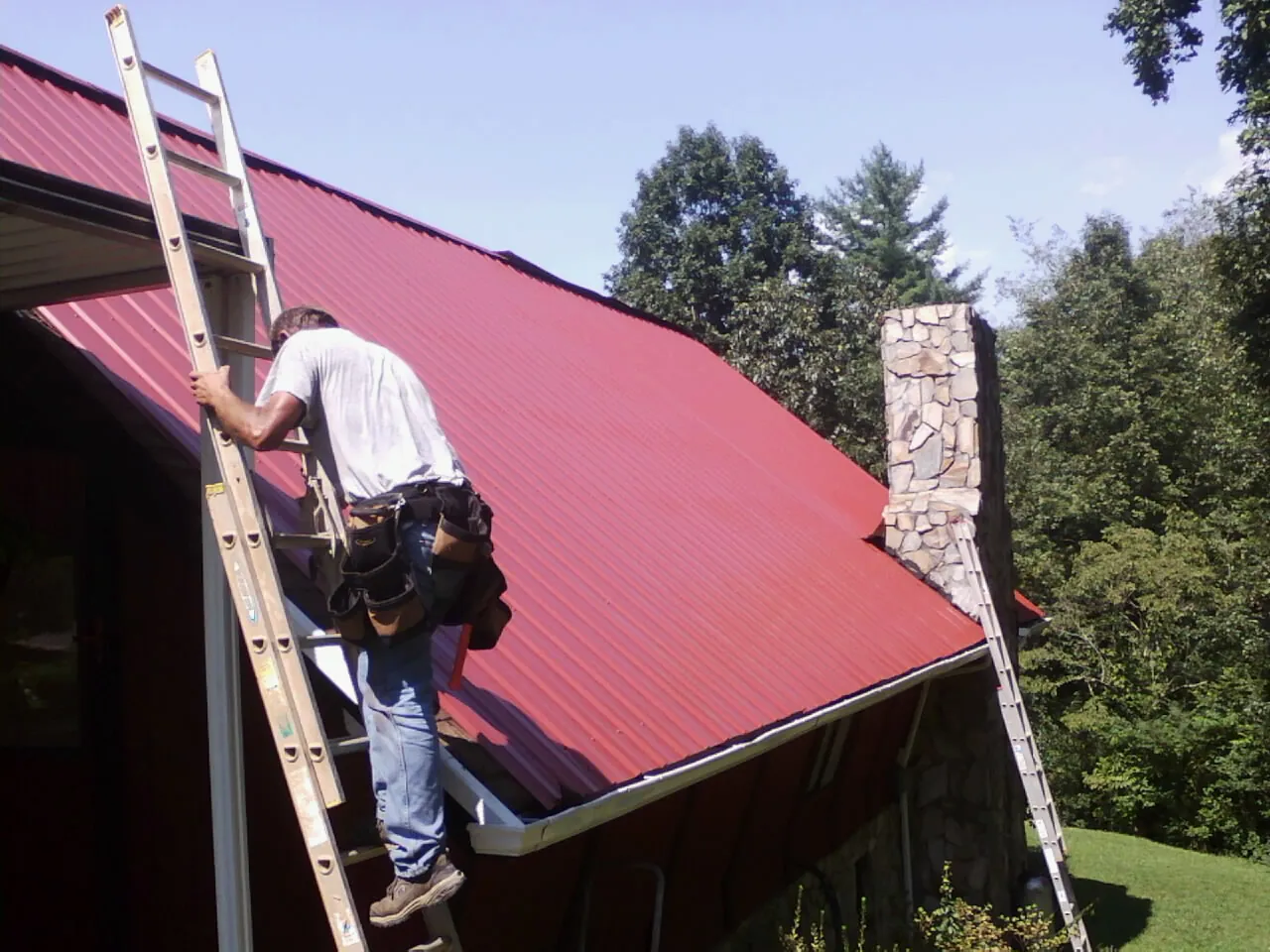 Roofer on a ladder beside a red metal roof and stone chimney — Soffit & Fascia Repair by Roofing Brothers in Pleasant Grove